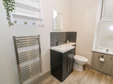 A bathroom with a black vanity unit with sink a toilet a heated towel rail a small white shelf with hooks and a window at Emerald Suite in Barmouth