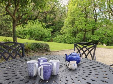An outdoor metal table with four patterned mugs a blue teapot and a small jug with trees and grass in the background at Glyn Yr Efail in Moylegrove nr Newport and Cardigan