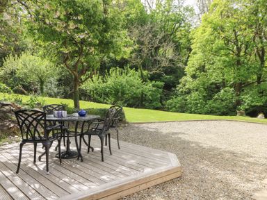 An outdoor patio with a metal table and chairs on a wooden deck surrounded by trees and a gravel area at Glyn Yr Efail in Moylegrove nr Newport and Cardigan