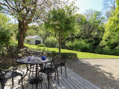 An outdoor patio with a round metal table and four chairs on a wooden deck with a green lawn and small white building in the background at Glyn Yr Efail in Moylegrove nr Newport and Cardigan