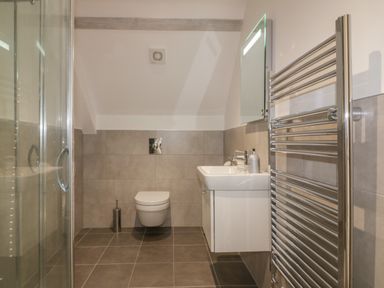 A bathroom with a wall-mounted toilet, a white sink with a mirror above, a shower enclosure, and a chrome towel rack at Stores & Supplies in Bodmin