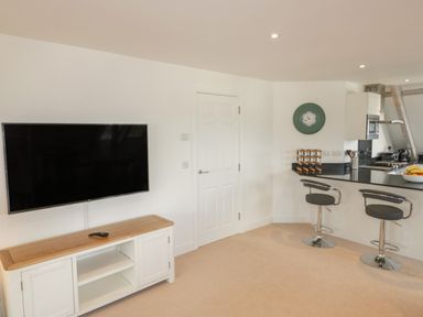 A room with a wall-mounted TV above a wooden cabinet a door and a kitchen counter with two bar stools at Stores & Supplies in Bodmin