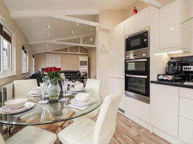A kitchen and dining area with a glass table set with white dishes and a vase of red roses at Sparrows Den Lodge in Winchelsea Beach