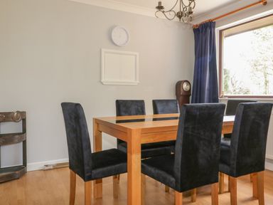 A dining room with a wooden table and six black chairs near a large window with blue curtains at Three Jays in Lanreath