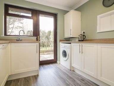 A kitchen with white cabinets wooden countertops a washing machine a kettle and a door leading outside at Three Jays in Lanreath