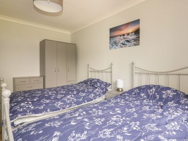 A bedroom with two single beds with blue floral bedding a wardrobe chest of drawers bedside table with lamp and ocean sunset artwork on the wall at Three Jays in Lanreath