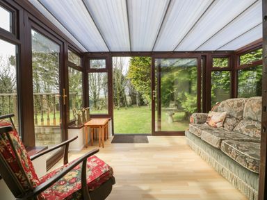 A conservatory with a floral sofa a chair with red patterned cushions two nesting tables and a garden view at Three Jays in Lanreath