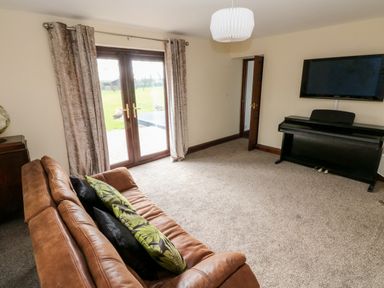 A room with a brown sofa with cushions a digital piano a wall mounted television curtains on double glass doors and a carpeted floor at Mountain Farm in Broad Haven