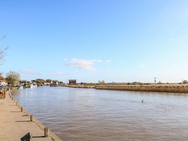 A river with boats docked on the left side and reeds on the right side under a blue sky at Foundry Cottage in Reedham