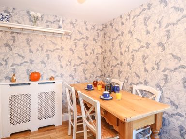 A dining room with a wooden table set with cups and glasses of juice and white chairs with a shelf and radiator cover against a patterned wall at Foundry Cottage in Reedham