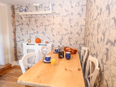 A dining area with a wooden table set with cups plates orange juice and a French press coffee pot with white chairs and patterned wallpaper at Foundry Cottage in Reedham