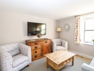 A living room with two patterned armchairs a wooden cabinet with flowers a wall clock and a coffee table at Foundry Cottage in Reedham