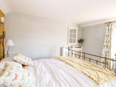 A bedroom with a metal bed frame white bedding patterned pillows a yellow patterned throw a white cabinet a lamp and patterned curtains at Foundry Cottage in Reedham