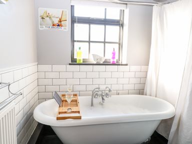 A bathroom with a white freestanding bathtub with bubbles and a wooden tray holding wine glasses under a window with bottles on the sill at Foundry Cottage in Reedham