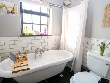 A bathroom with a clawfoot bathtub with bath tray holding glasses and a tablet near a window with bottles on the sill and a toilet with a plant on the tank at Foundry Cottage in Reedham
