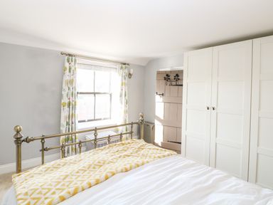 A bedroom with a metal bed frame a yellow patterned blanket white wardrobe a window with patterned curtains and a wooden door at Foundry Cottage in Reedham