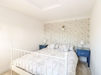 A bedroom with a white metal bed frame two blue bedside tables with lamps and patterned wallpaper at Foundry Cottage in Reedham