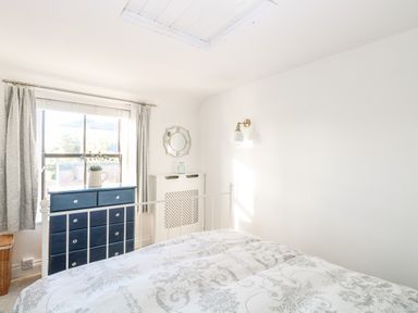 A bedroom with a bed a blue chest of drawers a window with curtains a wall mirror and a wall lamp at Foundry Cottage in Reedham