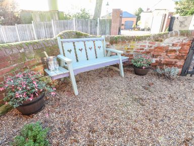 A wooden bench with heart cutouts beside a brick wall with two flower pots on gravel ground at Foundry Cottage in Reedham