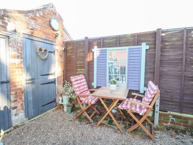 A small outdoor seating area with two wooden chairs with plaid cushions and a wooden table next to a brick wall and a fence with a decorative window frame at Foundry Cottage in Reedham