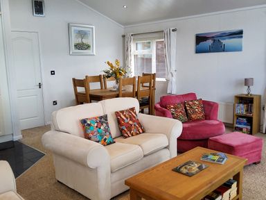 A living room with a beige sofa and colorful pillows a red armchair with matching ottoman a wooden coffee table and dining table with chairs at Lodge 11 in Tavistock