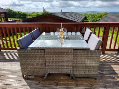 An outdoor patio dining set with four chairs and a glass top table with a bottle and four glasses on a wooden deck at Lodge 11 in Tavistock