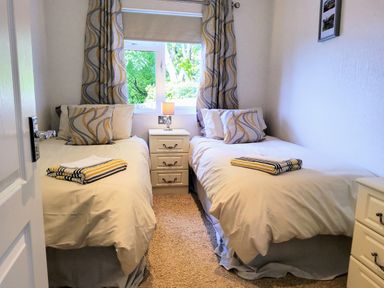 A bedroom with two single beds with patterned pillows and folded towels a lamp on a bedside table under a window with patterned curtains at Lodge 11 in Tavistock