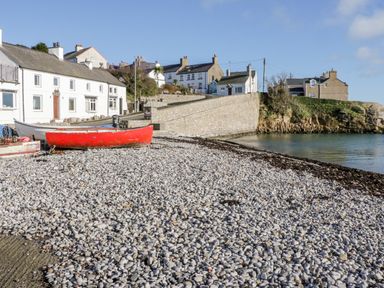 A pebble beach with a red boat near white houses and stone wall at Halcyon in Moelfre near Benllech