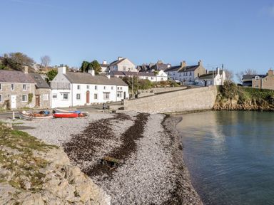 A pebble beach with boats near a stone wall and white houses at Halcyon in Moelfre near Benllech