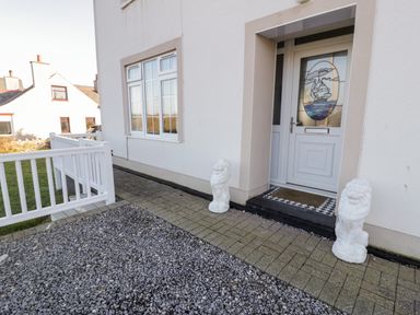The front entrance of a house with a white door featuring a stained glass image of a ship two white lion statues on either side of the door a stone pathway and a white railing fence at Halcyon Moelfre near Benllech