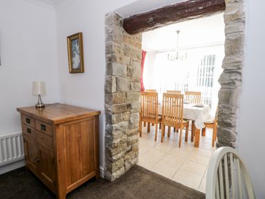 A wooden sideboard with lamp next to a stone archway leading to a dining area with wooden chairs and a table at Halcyon in Moelfre near Benllech
