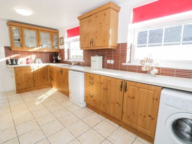 A kitchen with wooden cabinets dishwasher washing machine and red window blinds at Halcyon in Moelfre near Benllech