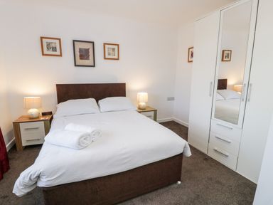 A bedroom with a double bed white bedding bedside tables lamps a wardrobe with mirror and framed pictures on the wall at Halcyon in Moelfre near Benllech