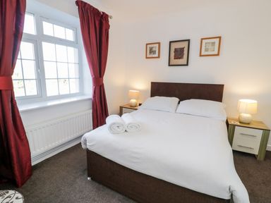 A bedroom with a double bed white bed linens bedside tables lamps red curtains and framed pictures on the wall at Halcyon in Moelfre near Benllech