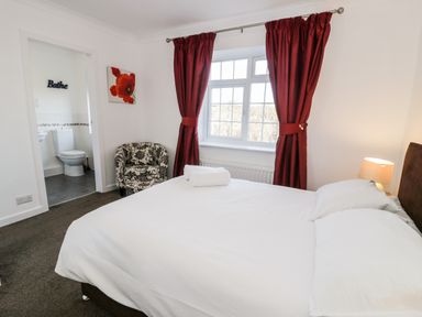 A bedroom with a bed white linens a chair red curtains and an ensuite bathroom at Halcyon in Moelfre near Benllech