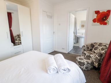 A bedroom with a white bed and two rolled towels a wardrobe with a mirror an armchair with a matching footstool a door leading to a bathroom and red curtains at Halcyon Moelfre near Benllech