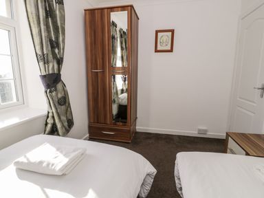 A bedroom with two single beds a wooden wardrobe with mirror white walls and a window with patterned curtains at Halcyon in Moelfre near Benllech