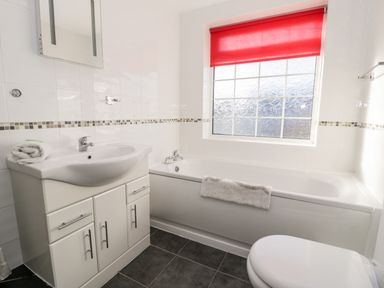 A bathroom with a sink, bathtub, toilet, and a window with a red blind at Halcyon Moelfre near Benllech