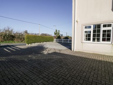 A paved driveway next to a house with a window and a hedge along a road at Halcyon in Moelfre near Benllech