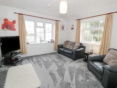 A living room with two black leather sofas cushions a television on a stand and windows with curtains at Halcyon Annexe in Moelfre near Benllech
