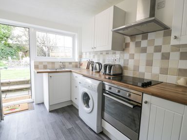 A kitchen with white cabinets a washing machine oven toaster kettle and door leading to garden at Halcyon Annexe Moelfre near Benllech