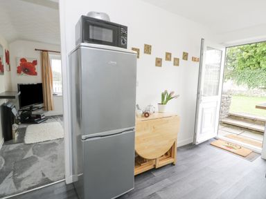 A kitchen area with a refrigerator and microwave next to a wooden drop-leaf table and an open door leading outside at Halcyon Annexe Moelfre near Benllech