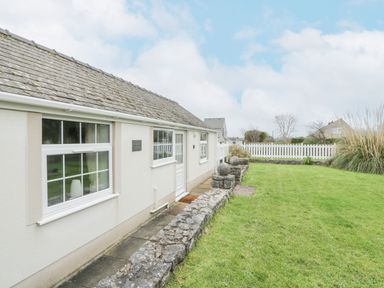 A white single-story house with a stone roof and a small garden with stone edging and green grass at Halcyon Annexe in Moelfre near Benllech