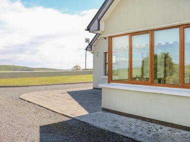 An exterior view of a building with large windows and a paved area next to a gravel driveway with grass in the background at Dooneen near Castletownshend County Cork