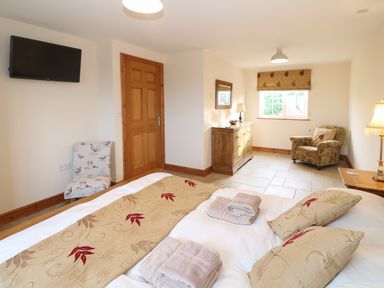 A bedroom with a bed with patterned pillows and folded towels a wooden dresser an armchair a floral chair a wall-mounted television and a window at Bryn Eiddon in Aberdaron