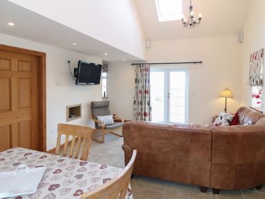 A living room with a brown leather sofa a wooden chair a dining table with a heart patterned tablecloth a wall-mounted television and double doors with curtains at Bryn Eiddon in Aberdaron