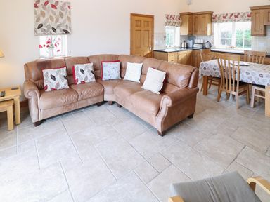 An open living room and kitchen with a brown leather corner sofa with cushions a wooden dining table with chairs and kitchen cabinets at Bryn Eiddon in Aberdaron