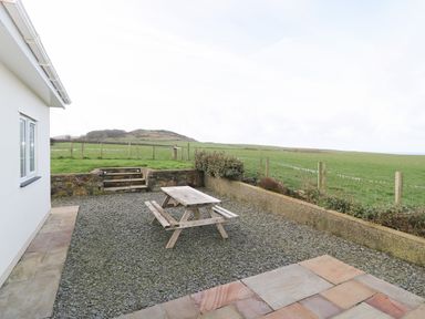 An outdoor patio area with a wooden picnic table and gravel ground near a white building and fenced grassy fields at Bryn Eiddon in Aberdaron