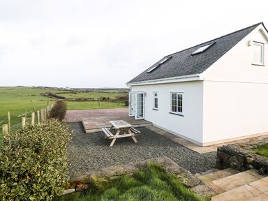 An outdoor area with a white house with skylights and windows a wooden picnic table gravel ground and green fields at Bryn Eiddon in Aberdaron
