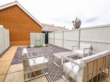 An outdoor patio with white cushioned chairs and a table on a patterned rug next to a fenced yard at 5 Styleman Road in Hunstanton